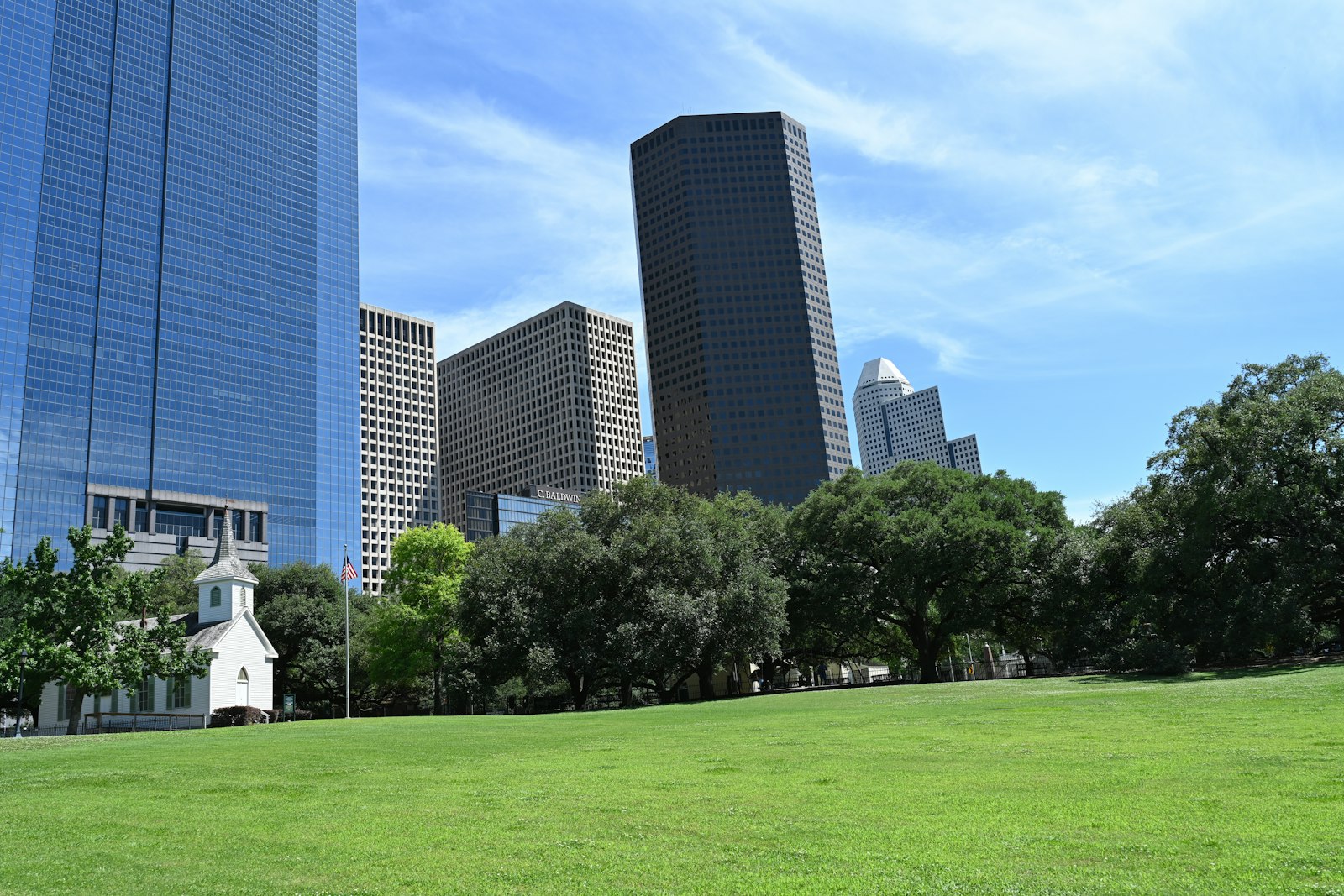 A small white historic church in a downtown Houston park with towers behind it